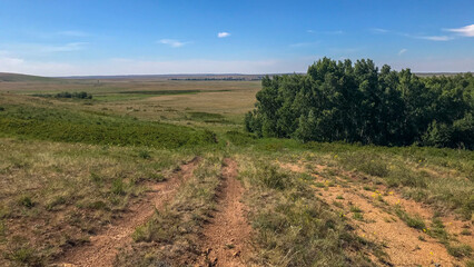 Expansive open prairie, with sparse trees and greenery scattered across it under a blue sky. In front is an off-road trail leading to distant hills. The landscape exudes a sense of tranquility.