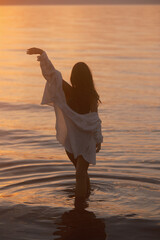 A woman in a green palm-print bikini and an unbuttoned white shirt stands in the water at dusk,...