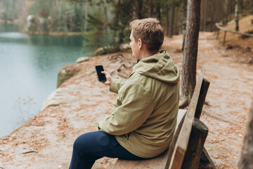 Lonely 30s blonde man with phone is sitting on the bench of a forest lake. Autumn landscape, calmness, tranquility, travel concept. A man on the bank of a lake is enjoying the nature.
