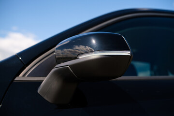 Close-up of a black car’s side mirror with an integrated turn signal. Glossy surface reflecting...