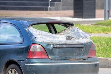 Old car with broken rear window covered with plastic film, parked on the side of a city road, realistic photo style, daylight outside, vehicle damage concept