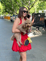 Young woman in red dress holding a poodle and smartphone walking on a Berlin city street

