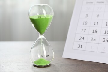 Deadline. Hourglass with flowing sand and calendar on wooden table indoors, closeup