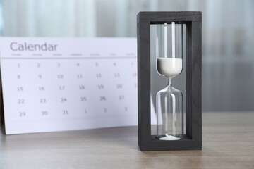 Deadline. Hourglass with flowing sand and calendar on wooden table indoors