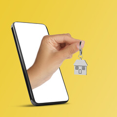 Woman's hand with keychain in shape of house sticking out from smartphone on yellow background....
