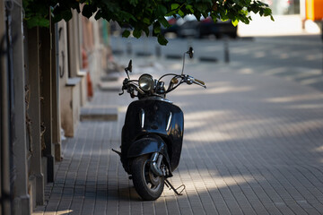 vintage black scooter parked on quiet european street under tree shade urban retro moped...