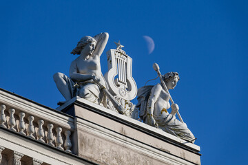 Naklejka premium classical sculptures on historic building rooftop with moon in background under clear blue sky in riga latvia european architecture detail art history rooftop decoration cultural heritage travel