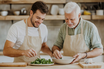 Young man and senior man preparing salad together in a cozy kitchen with natural wood elements. International Day of Families, Global Day of Parents, International Children Day