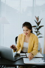 Asian accountant is calculating financial data for a business using a calculator and laptop in her...