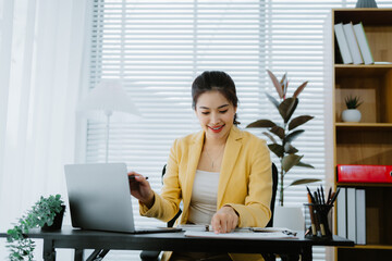 Asian accountant is calculating financial data for a business using a calculator and laptop in her...