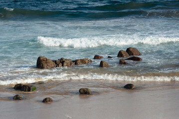 Dark rocks on the seashore of a beautiful beach. Waves breaking on the sand, paradise.