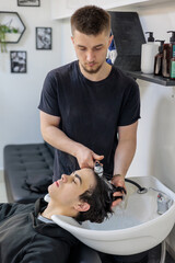 Teenager Getting Hair Washed at Modern Barbershop.  Male Barber Washing Client’s Hair at Sink in...