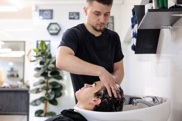 Teenager Getting Hair Washed at Modern Barbershop.  Male Barber Washing Client’s Hair at Sink in...