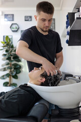 Teenager Getting Hair Washed at Modern Barbershop.  Male Barber Washing Client’s Hair at Sink in...