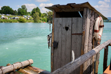 A weathered wooden outhouse with a heart-shaped cutout stands on a floating raft over turquoise...