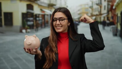 Hispanic woman smiling and flexing arm while holding a piggy bank on a bustling street showcasing strength and finance. - Powered by Adobe