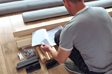 Focused man looking at instructions for self assembly, connects details of new bed in bedroom....