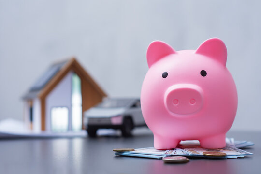 Pink piggy bank with US dollar and coins next to a miniature eco house with solar panels and an electric car model. Concept for financial goals, saving and sustainable living.