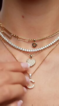 close-up static video on a woman's neck showcasing a layered stack of elegant diamond and gold necklaces including tennis, paperclip, charm, heart, initial 'c' pendant, with a hand adjusting the