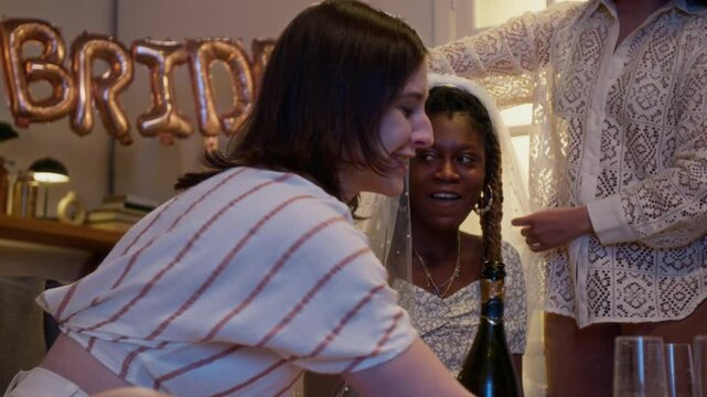 Group of best friends preparing beverages for bachelorette party. One of them putting veil on head of bride, they starting drinking champagne together