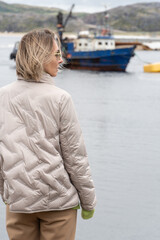 Fototapeta premium Thoughtful woman in a beige jacket and green sweater holding sunglasses while standing near a calm bay with an old blue fishing boat in the background