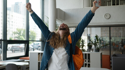 Caucasian student guy man male yelling celebrating winning with mobile phone in university excited...