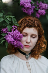 Young pretty caucasian woman in romantic outfit walking in blooming spring garden. Artistic outdoor photo of beautiful red-haired girl posing in fragrant lilac blossom.