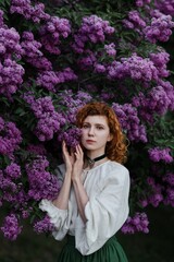 Young pretty caucasian woman in romantic outfit walking in blooming spring garden. Artistic outdoor photo of beautiful red-haired girl posing in fragrant lilac blossom.