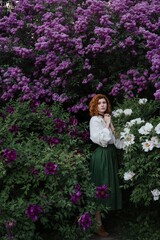 Young pretty caucasian woman in romantic outfit walking in blooming spring garden. Artistic outdoor photo of beautiful red-haired girl posing in fragrant lilac blossom.