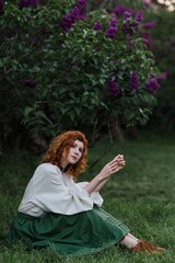Young pretty caucasian woman in romantic outfit walking in blooming spring garden. Artistic outdoor photo of beautiful red-haired girl posing in fragrant lilac blossom.