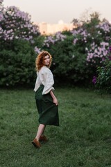 Young pretty caucasian woman in romantic outfit walking in blooming spring garden. Artistic outdoor photo of beautiful red-haired girl posing in fragrant lilac blossom.