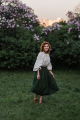 Young pretty caucasian woman in romantic outfit walking in blooming spring garden. Artistic outdoor photo of beautiful red-haired girl posing in fragrant lilac blossom.