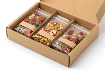 Box of multiple transparent sachets of granola, each sealed and labeled. Sachets filled with crunchy mix of oats, nuts, and fruit. Arranged neatly inside a brown cardboard box on a clean surface. Flat
