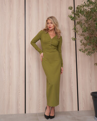 Blonde woman in an elegant green dress poses against a light wooden wall, hand on hip, with a...