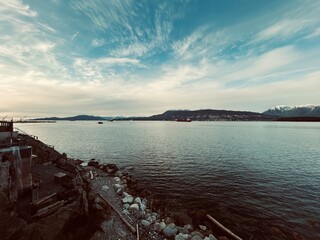 Coastal view with calm ocean water, rocky shoreline, mountains in the background, and a colorful sky during late afternoon.