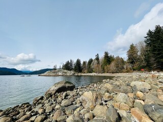 A peaceful rocky shoreline meets calm waters under a bright sky, with a backdrop of forested hills...