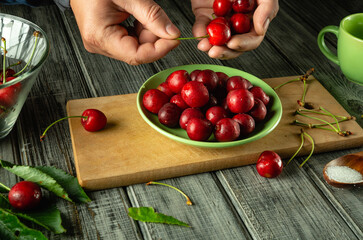 Hands are seen carefully sorting through ripe cherries in a green bowl on a wooden table. Leaves and utensils are scattered around, creating a fresh kitchen atmosphere