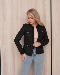 Blonde woman in black textured jacket and light jeans, standing against a light wood panel wall....