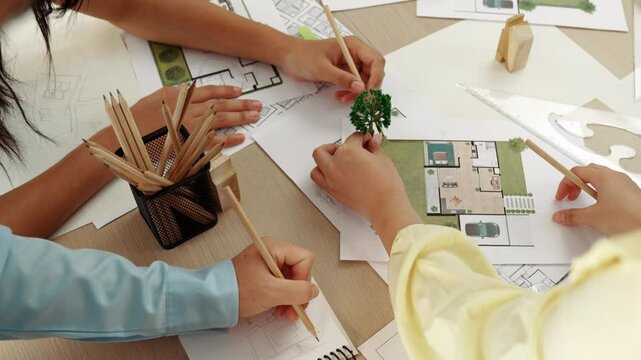Architect and engineer use tree model discuss about building blueprint on table with architectural document, measurement tools and wooden block scatter around at modern office. Closeup. Delineation.