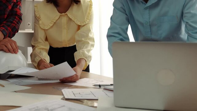 Architect and safety engineer discussion about building blueprint displaying on laptop on table with architectural document and wooden block scatter around at modern office. Closeup. Delineation.