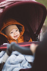 Smiling baby in a stroller wearing a striped hat and orange jacket on a chilly day