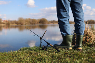 Cropped photo of fisherman's legs in rubber boots with rod, spinning reel on river bank. Unrecognisable man catching fish on lake, pond. Fishing for pike, perch on beach. Fishing day concept.