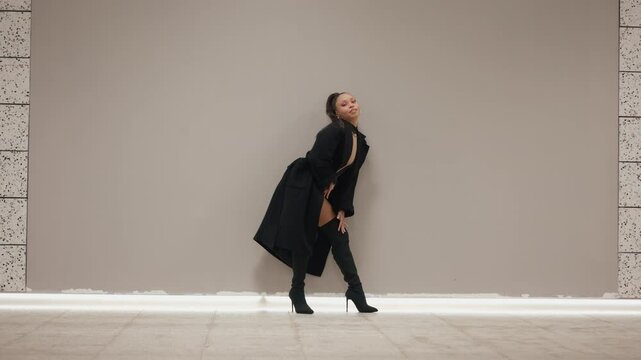 Confident woman fashion model dancing seductively and looking at camera in futuristic design underground subway hall. Female in black bodysuit, thigh-high boots and coat, concrete wall