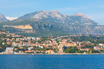 Fototapeta premium Herceg Novi town on the coast of Kotor Bay in Montenegro. Coastal town stretches along the shoreline, nestled at the base of sunlit mountains under a clear blue sky