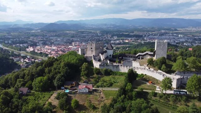 stunning aerial view of Celje Old Castle Celjski Stari Grad perched on a hill, surrounded by lush green forests. The castle overlooks the city of Celje, with the Savinja River winding through, and