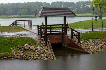 A traditional wooden water gate with a small pavilion sits by a calm river, surrounded by green...