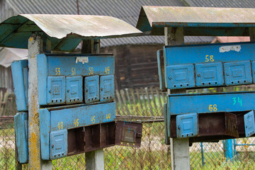 Rusty, faded blue communal mailboxes with numbered compartments stand on concrete posts, with old...