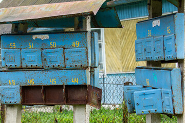 Rusty, faded blue communal mailboxes with numbered compartments stand on concrete posts, with old...