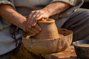 Skilled hands of a potter, covered in clay, expertly shape a pot on a traditional rustic wheel, embodying ancient craftsmanship and the art of pottery creation