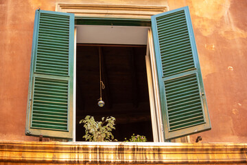An open window with vibrant green louvered shutters reveals a glimpse of an indoor plant and a hanging light, set against a warm, terracotta-colored building facade